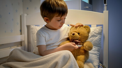 Portrait of cute sick boy measuring temperature to his toy teddy bear with digital thermometer....