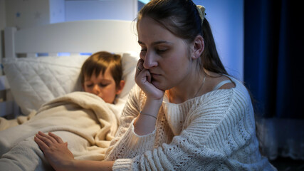 Upset, sad and stressed mother sitting next to bed of her sleeping child at night. Concept of...