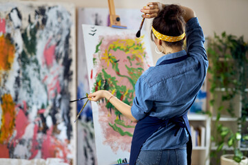 Back view of talented female artist holding paintbrushes with her hands in paint while thinking of...