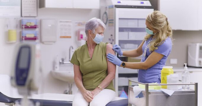 A Wide Shot Of An American Female Medical Nurse Administering A Covid-19, SARS-CoV-2 Vaccine Injection With A Needle and Syringe, To An Elderly Woman. Wearing Safety Gloves And Protective Mask
