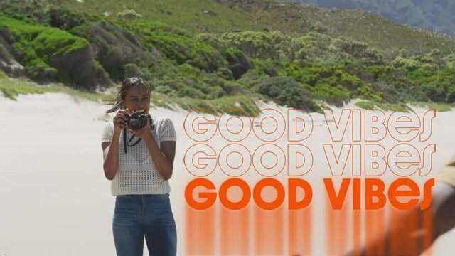 Animation of the words good vibes written in orange over woman on beach taking photos of male friend