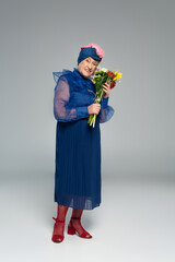 smiling elderly woman in blue dress and turban holding bouquet of flowers on grey