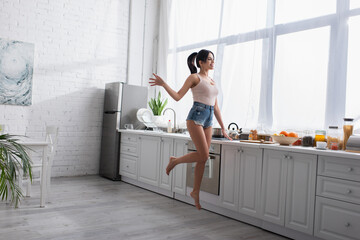 full length of cheerful young woman jumping near fruits in kitchen