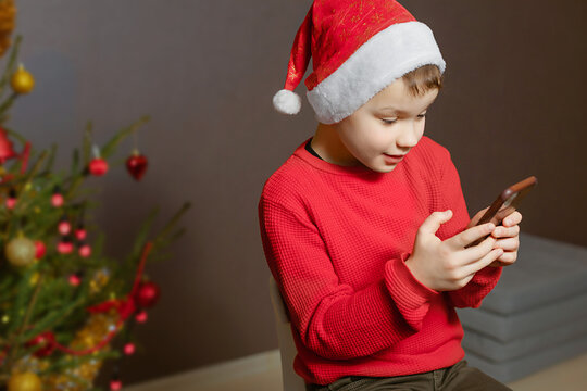 A boy in a red sweater and a stanta claus hat with a smartphone in his hands is typing a message indoors.