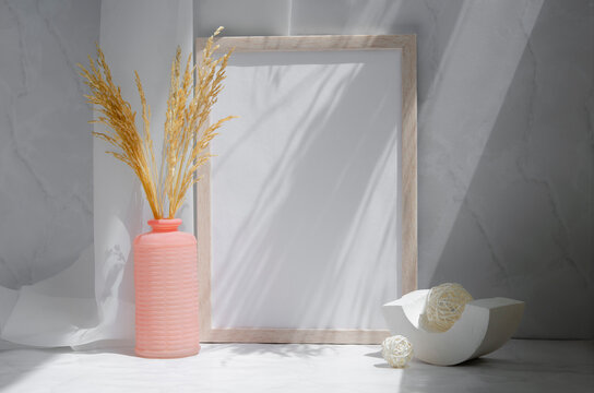 Empty picture frame on gray background. Interior design concept. Indoor modern still life. White blank wooden frame mockup, vase with dry grass, geometric shapes on the table.	