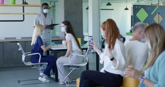 Patients sitting in queue in hospital waiting for mass immunization vaccination, professional female doctor and african male assistant at work confidently doing mass vaccination.focus on brunette
