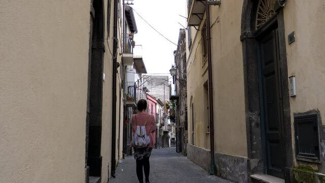 Rear view of young tourist woman with backpack walking narrow side street in Italian town Randazzo in Catania province, Italy.