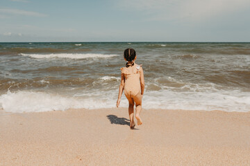 Fototapeta premium Six year girl in yellow swimsuit on the beach, people from behind. hot summer day.