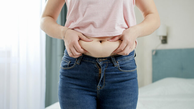 CLoseup of young woman in jeans holding fat fold on her belly. Concept of excessive weight, obese female, dieting and overweight problems