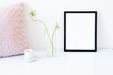 Blank black frame mockup. Small fresh flowers of white garlic (allium neapolitanum) in glass vase. Cup of coffee, pink embroidered pillow on glossy white table. White background