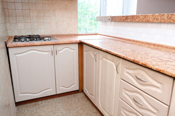 Interior design of modern narrow smart compact kitchen with white contemporary furniture, beige ceramic tiles on wall, gas stove and window view.