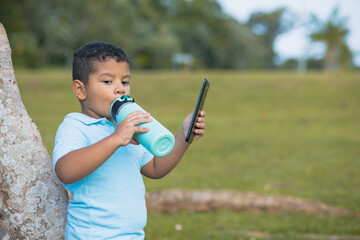 Latino boy leaning on the tree drinking water from a bottle