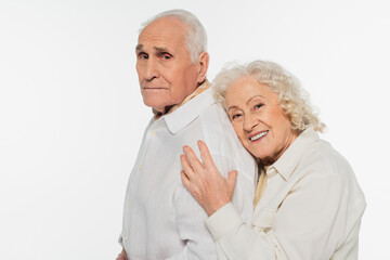happy elderly woman gently hugging back of husband and looking at camera isolated on white
