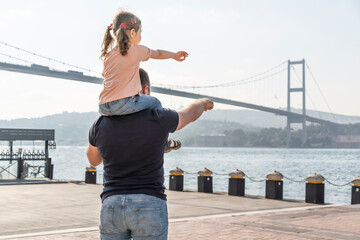 Little cute girl seat on father's shoulders in front of Bosphorus bridge (aka: 15 July Martyrs Bridge Turkish: 15 Temmuz Sehitler Koprusu) in Ortakoy, Istanbul, Turkey. Man with daughter.