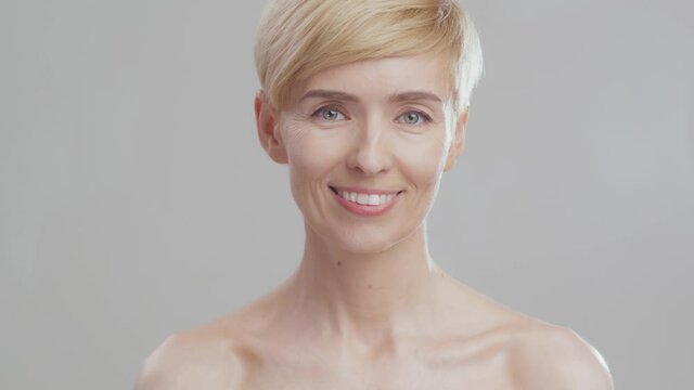 Gorgeous middle aged lady smiling to camera, posing with bare shoulders over grey studio background
