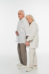 happy elderly woman gently hugging back of husband with holding hands and looking at camera on white