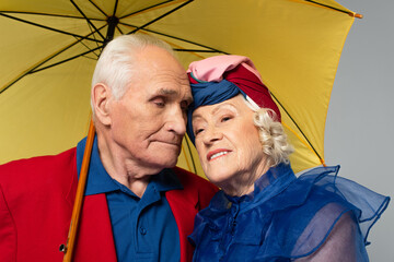 elderly man with yellow umbrella hugging wife in blue dress and turban isolated on grey