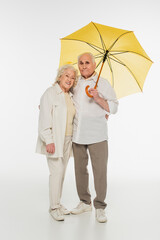 smiling elderly couple in casual clothes standing with yellow umbrella on white