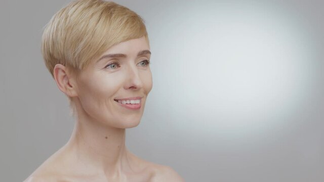 Semi profile portrait of happy middle aged blonde lady looking aside at empty space and smiling, grey background, mockup