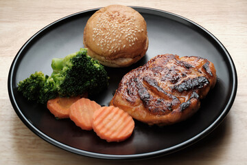 home made fried spicy chicken steak served with burger bread, sliced carrots, and pieces of broccolis in black dish on wooden table
