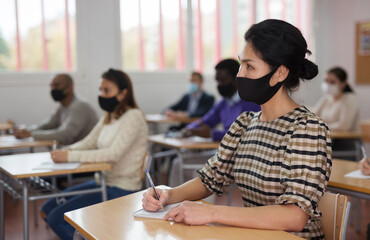 Portrait of focused female in protective face mask sitting at desk studying in classroom with colleagues