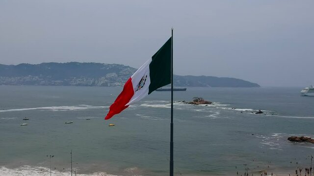 Acercamiento a&eacute;reo a bandera de M&eacute;xico ondeando con el viento con el mar y rocas de fondo