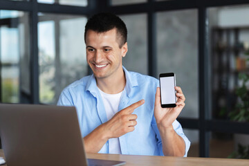 Attractive caucasian young man shows a mobile phone with a blank screen in laptop computer screen, video conference