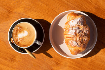 A cup of coffee and a croissant on a plate in the morning light on the table.