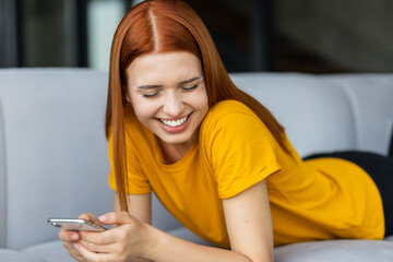 Smiling happy young woman with red hair texting on mobile phone lying on the couch at home