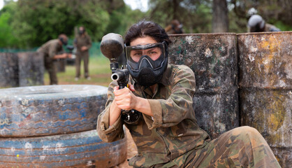 Fototapeta premium Female paintball player in camouflage and mask aiming with gun in shootout outdoors