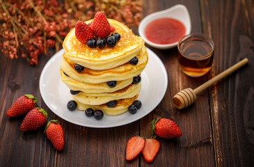 Sweet food. Stack of delicious pancakes with blueberries, Strawberry and Strawberry jam, fork rests in white plate on blur wooden background.