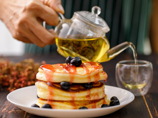 Close up hand pour the tea into the glass. Blur foreground pancakes with strawberry jam, decorated...