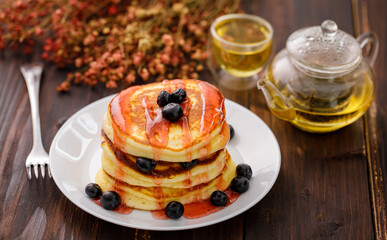 Sweet food. Stack of delicious pancakes with fork rests blueberries and honey, teapot in white plate on blur wooden background.