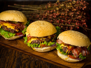 Three hamburgers with beef burger cutlet, fried onion, spinach, ketchup sauce and cheese, served on wood chopping board over dark wooden background.