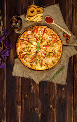 Close up on tasty homemade seafood pizza on sackcloth with dried purple flowers, cold beverage, Fried onions, potatoes, tomatoes and ketchup on wooden table. Food and Beverage Concept.