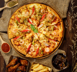 Close up on tasty homemade seafood pizza on sackcloth with dried plants, cold beverage, fried onions, potatoes, chicken, oregano and ketchup on wooden table. Food and Beverage Concept.
