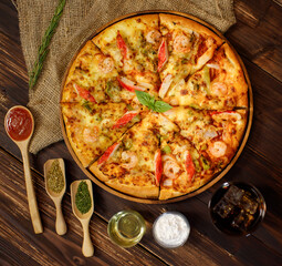 Close up on tasty homemade seafood pizza on sackcloth with dried plants, cold beverage, fried onions, potatoes, chicken, oregano and ketchup on wooden table. Food and Beverage Concept.