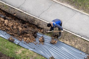 A worker digging a ditch along a pavement in summer.