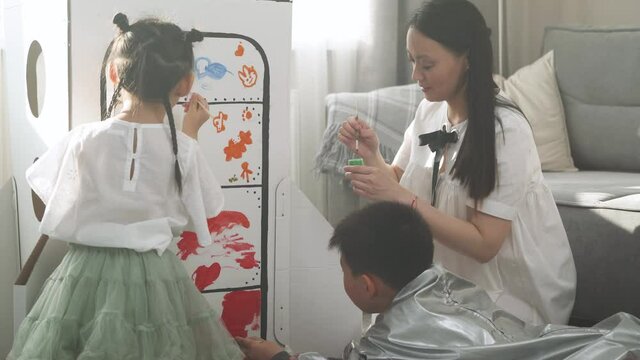 An Asian female with kids play in the living room at home, a boy in an astronaut costume sitting on the floor with her mother and sister, children together with their mother paint on a cardboard model
