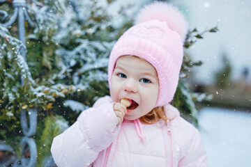 Portrait of little toddler girl walking outdoors in winter. Cute toddler eating sweet lollypop candy. Child having fun on cold snow day. Wearing warm baby pink clothes and hat with bobbles.