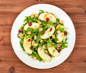 Fresh green apple slice salad with rocket leaf, oat granular, dried cranberry, and cashew nut placed in white dish on the dark wooded table