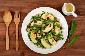 Fresh green apple slice salad with rocket leaf, oat granular, dried cranberry, and cashew nut placed in white dish on the dark wooded table