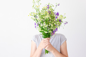 A portrait of a young girl in gray clothes who hid her face behind a bouquet of purple and yellow wildflowers and grass against a white wall.