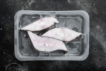 Frozen Greenland halibut steaks vacuum pack, on black dark stone table background, top view flat lay