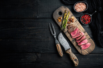 Juicy steak medium rare beef Filet Mignon or Eye Fillet, with onion and asparagus, on wooden serving board, with meat knife and fork, on black wooden table background, top view flat lay - obrazy, fototapety, plakaty