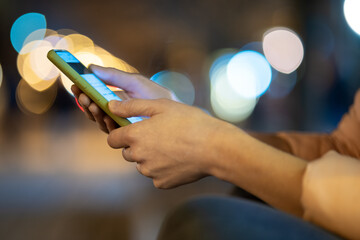 Closeup of female hands holding mobile phone browsing internet on city street with blurred lights at night.