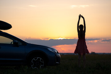 Naklejka na ściany i meble Dark silhouette of woman driver standing near her car on grassy field enjoying view of bright sunset. Young female relaxing during road trip beside SUV vehicle.