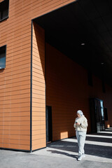 Vertical wide angle portrait of modern Middle-Eastern woman using smartphone in city lit by sunlight and wearing headphones, copy space