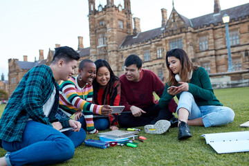 Group of young university students hanging out sitting on grass studying and using devices