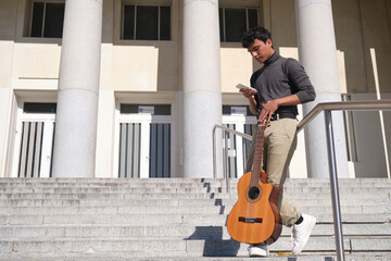 Latin young man chatting on his smartphone with a guitar standing on stairs. Waiting for something.